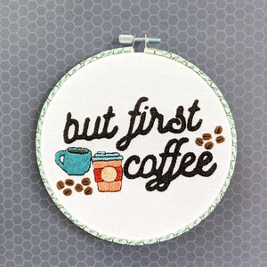 Puede incluir: Arte de bordado en forma de aro con el texto "but first coffee" en fuente cursiva negra. El dise&ntilde;o incluye dos tazas de caf&eacute;, una azul y una marr&oacute;n con una tapa roja, y granos de caf&eacute; marrones.