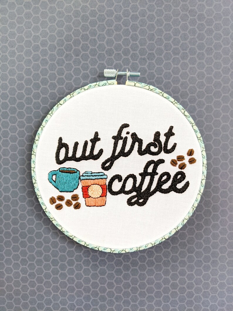 Puede incluir: Arte de bordado en forma de aro con el texto "but first coffee" en fuente cursiva negra. El dise&ntilde;o incluye dos tazas de caf&eacute;, una azul y una marr&oacute;n con una tapa roja, y granos de caf&eacute; marrones.