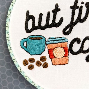 Puede incluir: Arte de bordado en aro con el texto "but first, coffee" en cursiva negra. El dise&ntilde;o incluye dos tazas de caf&eacute;, una azul y una naranja, y granos de caf&eacute; marrones.