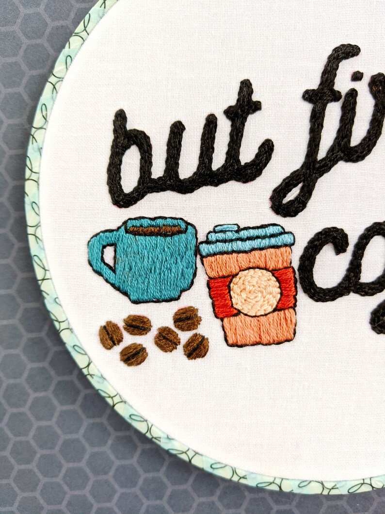 Puede incluir: Arte de bordado en aro con el texto "but first, coffee" en cursiva negra. El dise&ntilde;o incluye dos tazas de caf&eacute;, una azul y una naranja, y granos de caf&eacute; marrones.