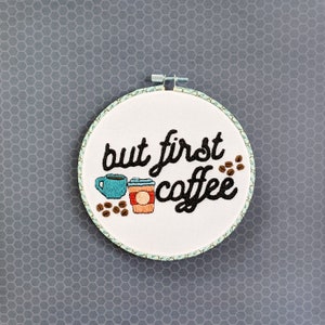 Puede incluir: Arte de bordado en aro con el texto "but first coffee" en letras cursivas negras. Dos tazas de caf&eacute; est&aacute;n bordadas debajo del texto, una azul y una roja.