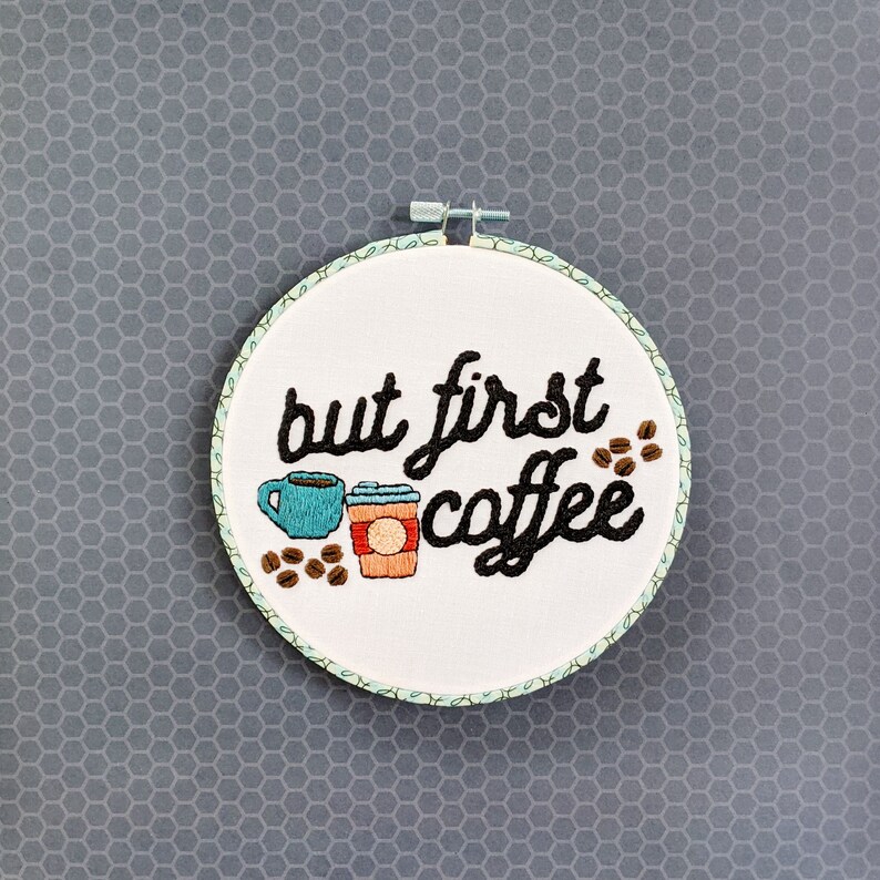Puede incluir: Arte de bordado en aro con el texto "but first coffee" en letras cursivas negras. Dos tazas de caf&eacute; est&aacute;n bordadas debajo del texto, una azul y una roja.