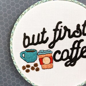 Puede incluir: Arte de bordado en aro con el texto "but first, coffee" en hilo negro. El dise&ntilde;o incluye una taza de caf&eacute; azul, un vaso de caf&eacute; para llevar marr&oacute;n y naranja, y granos de caf&eacute; marrones.