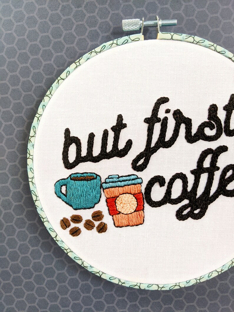 Puede incluir: Arte de bordado en aro con el texto "but first, coffee" en hilo negro. El dise&ntilde;o incluye una taza de caf&eacute; azul, un vaso de caf&eacute; para llevar marr&oacute;n y naranja, y granos de caf&eacute; marrones.