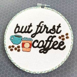 Puede incluir: Arte de bordado en aro con el texto "but first coffee" en fuente cursiva negra. El dise&ntilde;o incluye dos tazas de caf&eacute;, una azul y una naranja, y granos de caf&eacute; marrones.