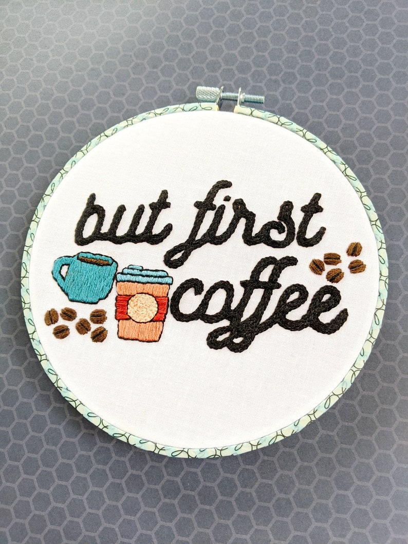 Puede incluir: Arte de bordado en aro con el texto "but first coffee" en fuente cursiva negra. El dise&ntilde;o incluye dos tazas de caf&eacute;, una azul y una naranja, y granos de caf&eacute; marrones.