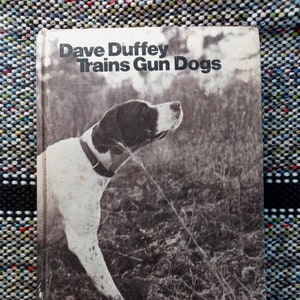 May include: A black and white photo of a book titled "Dave Duffey Trains Gun Dogs". The cover features a black and white photo of a dog standing in tall grass.