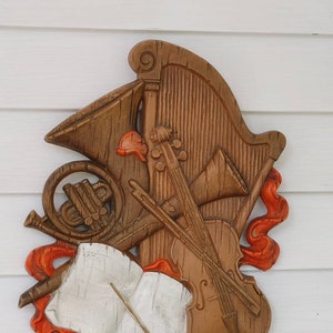 May include: A wooden wall hanging featuring musical instruments, including a harp, violin, and French horn. The instruments are brown with orange accents and a white sheet music element. The design is a decorative representation of music.