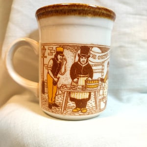 May include: White ceramic mug with a brown rim and handle. The mug features a sepia-toned illustration of a street scene with figures and the text "THE OYSTERS". The mug is a standard size for coffee or tea.