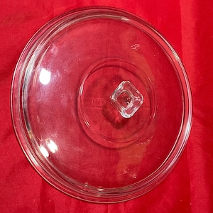 May include: A clear glass lid with a circular design. The lid features a raised rim and a square knob in the center. The glass is transparent, allowing a view of the red surface beneath.