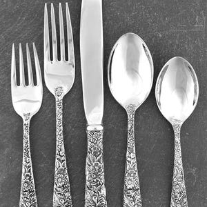 May include: A set of five silver-colored utensils: two forks, a knife, and two spoons. The handles are intricately decorated with floral patterns. The knife blade is smooth and reflective. The utensils are arranged vertically against a dark background.