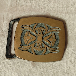 May include: A gold-toned belt buckle with a green, intricate design. The buckle has a rectangular shape with rounded corners.