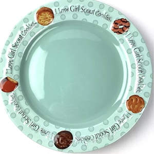 May include: A light blue ceramic plate with a white and green floral pattern. The plate has a rim with the text "I Love Girl Scout Cookies" repeated around the edge. There are images of Girl Scout cookies around the rim of the plate.