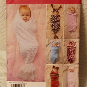May include: Simplicity sewing pattern number 1898A for making baby swaddles. The pattern includes instructions for seven different swaddles, each with a unique design and color scheme. The swaddles are shown in the image, with a baby in each one. The pattern is available in sizes XXS-XS, XXP-XP, and TTP-TP.