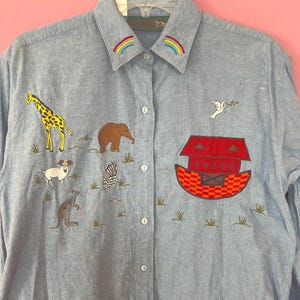 May include: A light blue denim shirt with embroidered animals and a rainbow on the collar. The animals include a giraffe, elephant, sheep, zebra, kangaroo, and a dove with an olive branch. The shirt also features a red and orange embroidered ark.