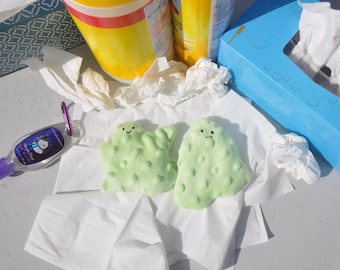 Può includere: Due giocattoli di peluche verdi a forma di fantasmi con facce sorridenti. I giocattoli sono seduti su una superficie di carta velina bianca. Ci sono altri oggetti sullo sfondo, tra cui una scatola di fazzoletti blu, un contenitore giallo e una bottiglia di disinfettante per le mani.