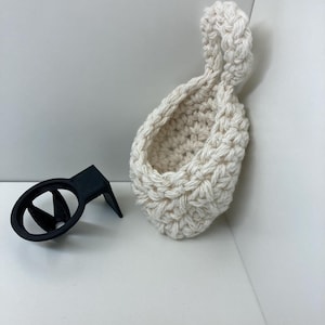 Puede incluir: Un soporte negro para teléfono de ventilación de coche, impreso en 3D, junto a una pequeña cesta de ganchillo blanquecina. La cesta tiene un lazo en la parte superior. Los artículos están sobre una superficie blanca.