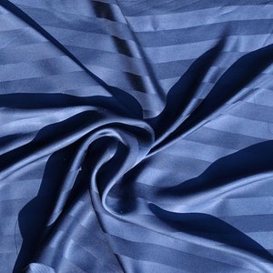 Op de afbeelding: Een close-up van een blauwe gestreepte stof met een gladde, zijdeachtige textuur. De stof is gevouwen en gedrapeerd, waardoor een zachte en elegante uitstraling ontstaat.