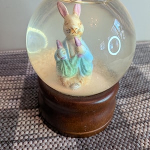 Puede incluir: Un globo de nieve con una figura de conejo con orejas rosas, vestido con un atuendo azul y verde y sosteniendo zanahorias moradas. El globo está sobre una base de madera marrón. El fondo es de color neutro.