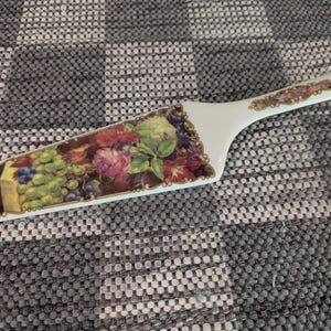 May include: White ceramic cake server with a decorative floral and grape design. The server features a wide, rectangular blade with a curved handle. The blade is adorned with a colorful illustration of grapes, flowers, and leaves, with gold trim.