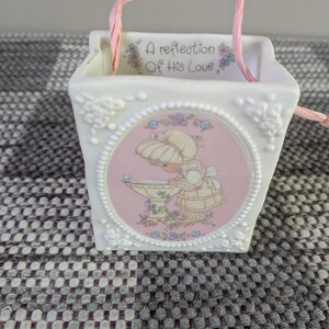 May include: White ceramic basket with a pink handle and a circular image of a girl. The image is surrounded by a white frame. The text "A reflection Of His Love" is printed on the top rim of the basket.