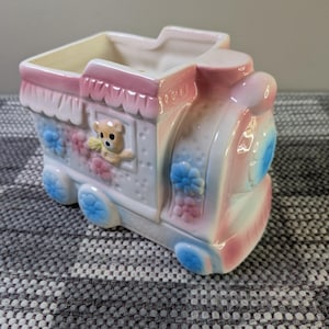May include: A ceramic train-shaped planter in pastel pink, blue, and white. The planter features a teddy bear accent and flower details. The train has blue wheels and a rectangular opening for plants.