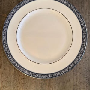 May include: A white ceramic plate with a blue and silver decorative border. The plate has a gold trim and a smooth, glossy surface. This elegant plate is suitable for formal dining.
