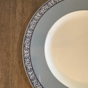 May include: A partially visible, round, white and blue plate with a decorative border. The plate has a light blue rim with a dark blue and white ornate pattern. The center of the plate is white. The plate is on a wooden surface.