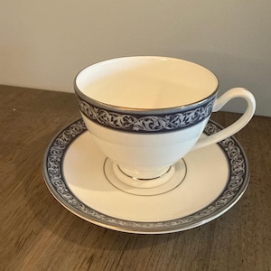 May include: White teacup and saucer set with a navy blue and gold decorative border. The teacup has a curved handle and sits on a matching saucer. The set is likely made of porcelain or fine china.