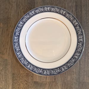 May include: A white plate with a blue and silver decorative border. The plate has a gold rim and a simple, elegant design. The plate is likely for serving food and is a part of a dinnerware set.