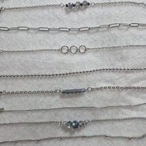 May include: A collection of ten silver-toned metal chains arranged horizontally. The chains feature various designs including simple links, ball chains, snake chains, and delicate strands accented with small iridescent beads and circular rings.