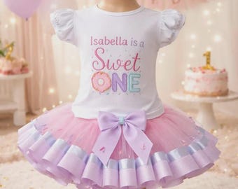 May include: A white t-shirt with the text "Isabella is a Sweet ONE" and a donut graphic, paired with a pink and lavender tutu skirt. The outfit is designed for a first birthday celebration.