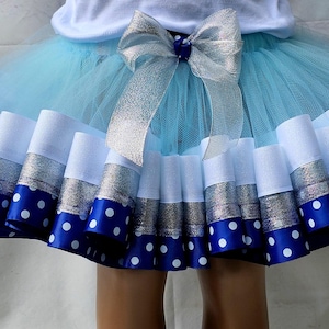 May include: A light blue tutu skirt with layers of tulle and decorative ribbons. The skirt features white, silver, and blue polka dot ribbons. A large silver bow adorns the waist. This skirt is suitable for dance or dress-up.