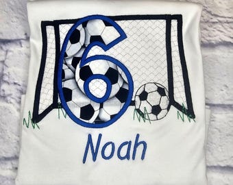 May include: White t-shirt with a blue number six made of soccer balls in a soccer goal. The name Noah is embroidered below the number.