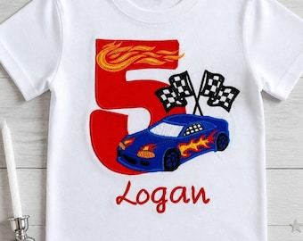 May include: A white short-sleeve t-shirt featuring an embroidered design of a large red number five with orange flames, a blue race car with yellow flames, two black and white checkered flags, and the name Logan embroidered in red script.
