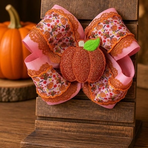 May include: A hair bow featuring pink ribbon with a pattern of orange pumpkins and turkeys, accented with orange lace trim. A glittery orange pumpkin applique with a green leaf sits in the center of the bow, which is displayed on a wooden stand.
