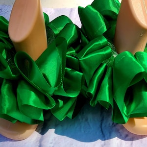 May include: A pair of green satin ankle cuffs with ruffles. The cuffs are on a pair of mannequin legs.