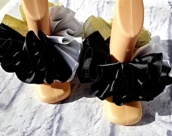 May include: A pair of decorative ankle cuffs with layered black, white, and gold ribbons. The cuffs are displayed on mannequin feet against a white brick wall background. The ribbons are ruffled and create a voluminous effect.
