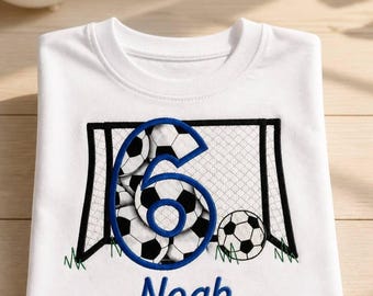 Puede incluir: Una camiseta blanca con un diseño bordado de temática futbolística. El gráfico incluye una portería de fútbol en blanco y negro, balones de fútbol y un gran número 6 azul. El nombre Noah está bordado en azul debajo del gráfico de la portería.