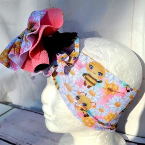 May include: A pink and white headband with a large pink bow and a bee print. The headband features a pattern of bees, flowers, and honeycomb.
