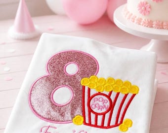 May include: A white t-shirt featuring an embroidered design of a large pink glittery number eight next to a red and white striped popcorn bucket with yellow popcorn. The name Emilia is embroidered in pink script below the graphic.