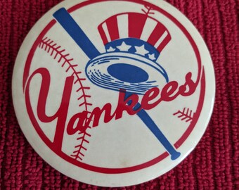 May include: A white button with a red, white, and blue baseball logo. The logo features a baseball bat, a baseball, and a hat with stars and stripes. The word "Yankees" is written in red and blue letters.