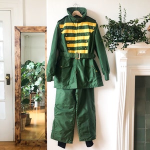 May include: A green ski suit with a yellow and gold striped jacket. The jacket has a belt and multiple pockets. The trousers are a solid green.