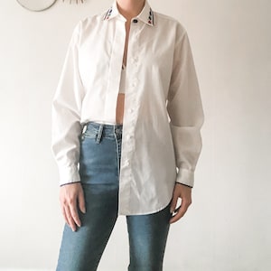 May include: A white long-sleeved button-down shirt with a colourful embroidered trim around the collar. The shirt is slightly oversized and has a relaxed fit.