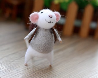 May include: A white felt mouse with pink ears and a gray sweater is wearing a brown felt backpack. The mouse is standing on a wooden surface.