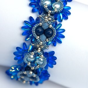 K&ouml;nnte beinhalten: Ein blaues Perlenarmband mit silbernen Akzenten und blauen und wei&szlig;en Perlen. Das Armband besteht aus kleinen, zarten Perlen und hat ein florales Design.
