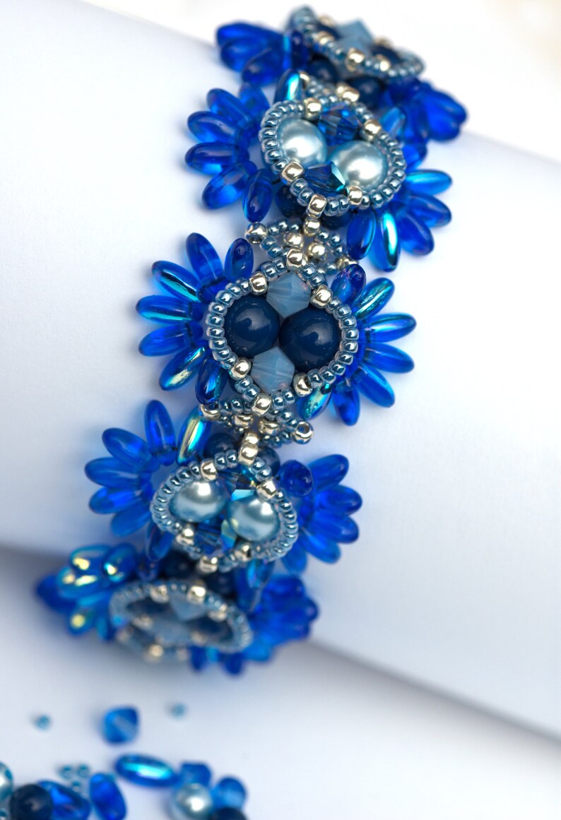 K&ouml;nnte beinhalten: Ein blaues Perlenarmband mit silbernen Akzenten und blauen und wei&szlig;en Perlen. Das Armband besteht aus kleinen, zarten Perlen und hat ein florales Design.