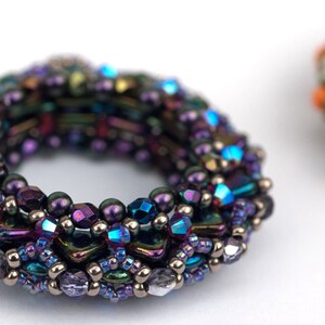 May include: A close-up of a purple, blue, and silver beaded bracelet. The bracelet is made of small, round beads and is in the shape of a ring. The bracelet is on a white surface.