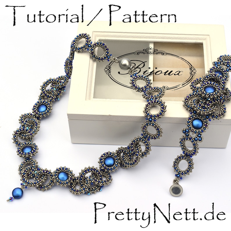 Może przedstawiać: Srebrny naszyjnik z niebieskimi koralikami i srebrnym zapięciem. Naszyjnik wykonany jest z małych, okrągłych koralik&oacute;w i charakteryzuje się wzorem splecionych k&oacute;ł. Naszyjnik jest prezentowany na białym pudełku z napisem "Bijoux". Na zdjęciu widoczny jest r&oacute;wnież tekst "PrettyNett.de".