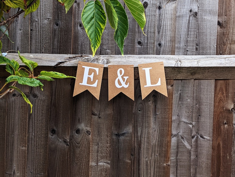 May include: A banner with the letters "E & L" in white on brown pennants. The banner is strung across a weathered wooden fence. Green leaves and branches are visible in the background, adding a natural touch.